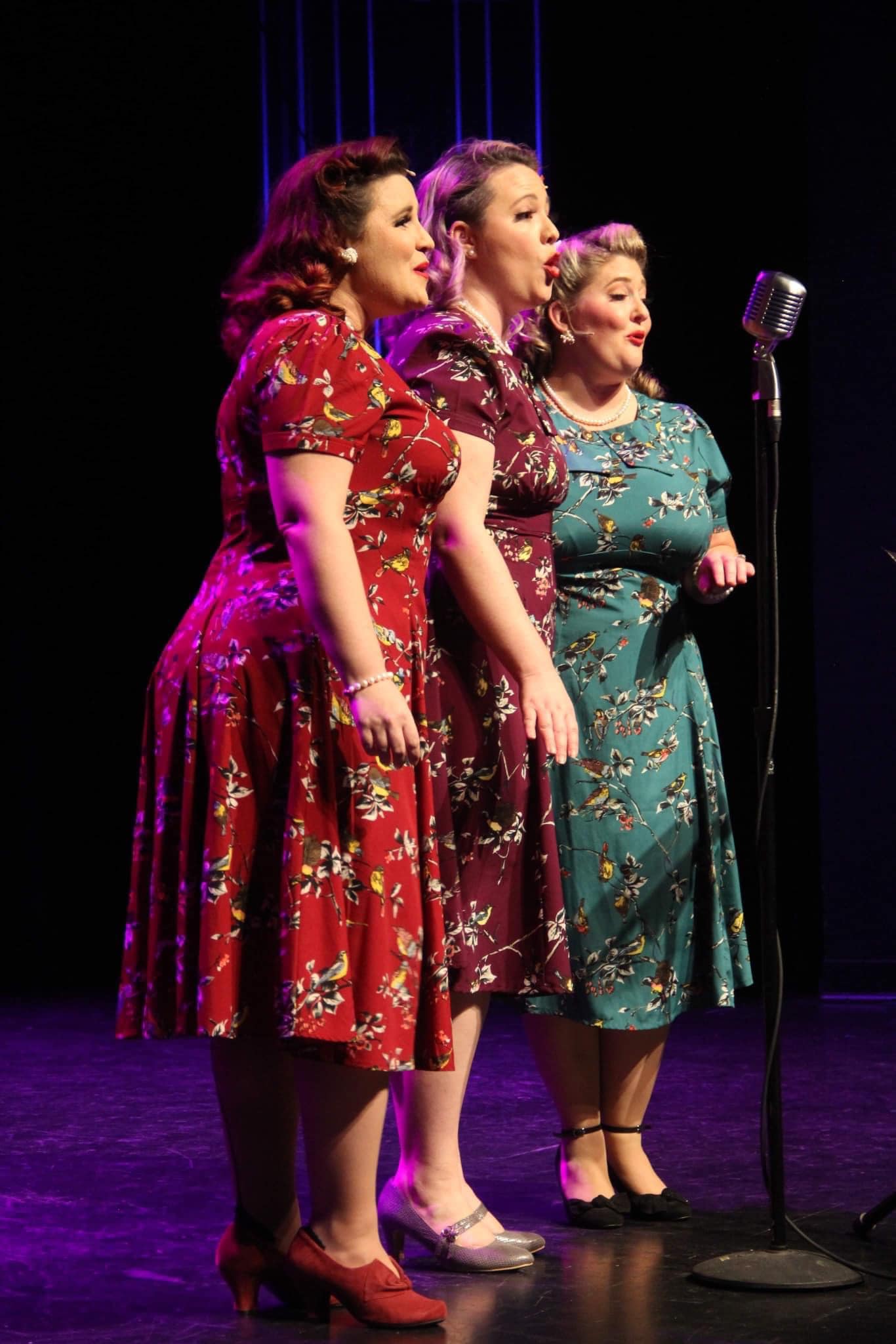 Three performers in vintage dresses singing at a microphone on stage